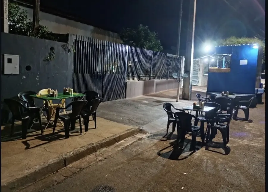 Mesas e cadeiras na área externa da Ferracini Lanches, à noite, ao lado do trailer de atendimento